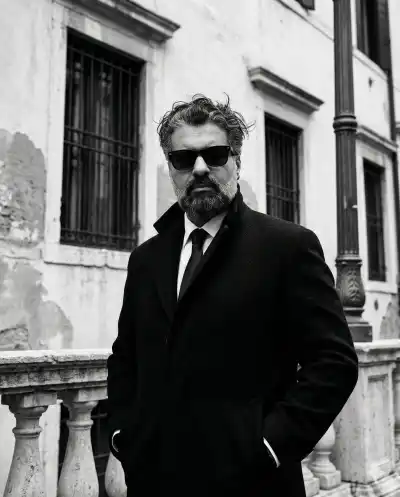 Black and white editorial fashion portrait of a handsome man with wavy tousled dark hair, wearing a heavy black tailored topcoat, white shirt, dark tie, and thick rectangular sunglasses